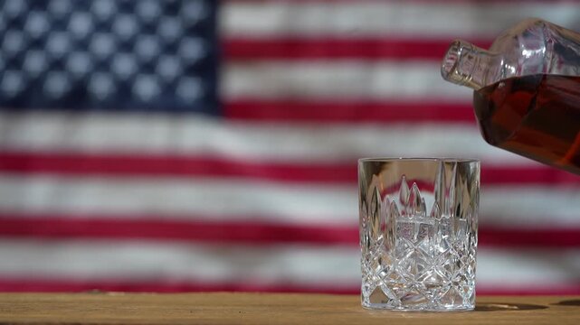 Pouring whiskey from bottle into glass on table against the backdrop of an American flag. Bourbon, an American whiskey, is often associated with the United States and is considered the national drink