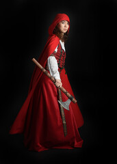 dramatic portrait of pretty young female brunette  model wearing a red hooded fairytale cloak and medieval historical gown. Holding a axe weapon. Isolated figure on dark studio background.