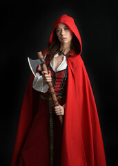 dramatic portrait of pretty young female brunette  model wearing a red hooded fairytale cloak and medieval historical gown. Holding a axe weapon. Isolated figure on dark studio background.