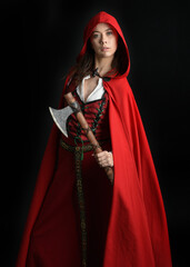 dramatic portrait of pretty young female brunette  model wearing a red hooded fairytale cloak and medieval historical gown. Holding a axe weapon. Isolated figure on dark studio background.