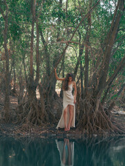 Young woman in a white dress stands barefoot by a forest lake, among tree roots and reflected in...