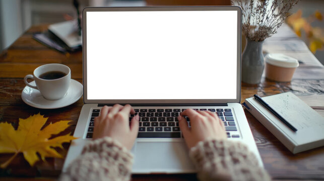 Person types on a laptop with a white screen, putting a cup of coffee, a book, a pen, a vase with dried flowers and a yellow maple leaf in front of him. Laptop mockup. Back to school. Cozy home office - Powered by Adobe