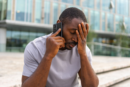 Sad tired lonely disappointed African man alone talking on smartphone on city street outdoor. Unhappy person making answering call by cellphone experiencing grief getting bad news. Stressed man
