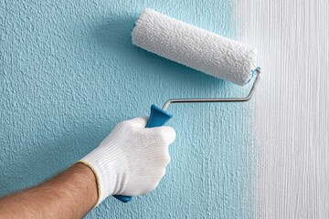 Obraz premium A hand in a white glove paints a textured wall light blue, leaving a clean white stripe of unpainted space.