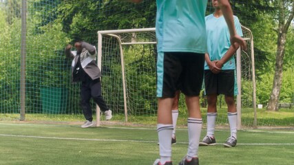 Slowmo of young soccer player lining up powerful kick toward goalpost while Black male coach catching and two diverse teammates observing, capturing focused moment in outdoor team training