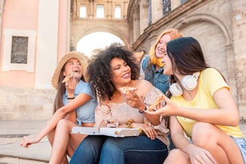 Four young cheerful multiethnic women having fun eating pizza outdoors in the city