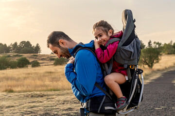 A happy father carrying his daughter in a child carrier backpack on a hiking trip. A smiling man and his child enjoying an outdoor adventure and an active family lifestyle in nature.