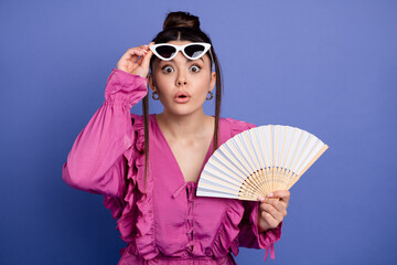Fashionable young woman in pink dress holding a handheld fan, looking surprised, posing against a...