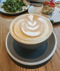 A cup of cappuccino with a decorative latte art design is served on a saucer on a wood table.