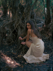 Young pensive woman in white dress sits and rests in forest among old tree roots.