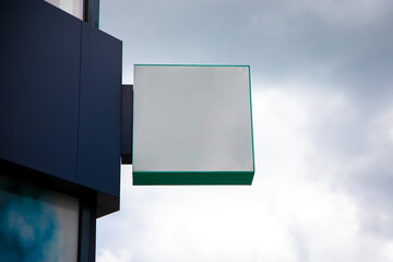 Empty square signboard mounted on a modern building facade with cloudy sky background, ready for...