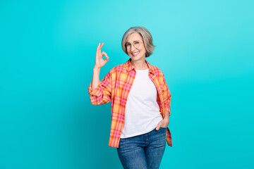 Smiling mature woman showing Ok gesture wearing casual clothes on bright turquoise background.