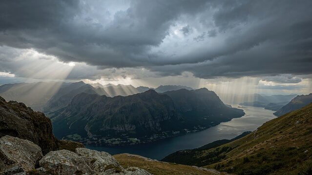 Ominous storm clouds hover above high mountains and a tranquil fjord, with intermittent rain and radiant light beams piercing the storm, visible from a hiking route to a mountain peak.