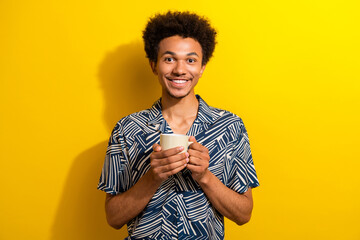 Young cheerful man holding white cup in front of yellow wall, wearing stylish patterned shirt,...
