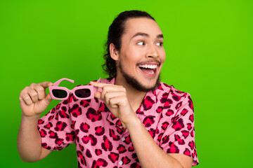Excited young man in stylish pink leopard print shirt holding sunglass on vibrant green background,...