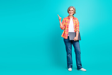 Smiling Mature Woman Pointing Upwards with Laptop in Trendy Casual Attire Against Bright Blue...