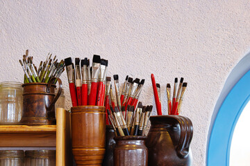A wooden shelf in rustic artist studio or creative workshop, with glass jars and clay jugs filled with paintbrushes 
