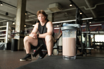 Protein shake in foreground and blurred athlete seated in gym, focusing on recovery. Workout at...