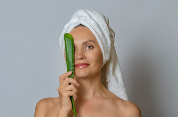 Woman wrapped in white towel covering one eye with aloe leaf and smiling. Gray studio background.