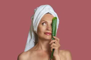 Woman wrapped in white towel covering one eye with aloe leaf and smiling. Pink studio background.