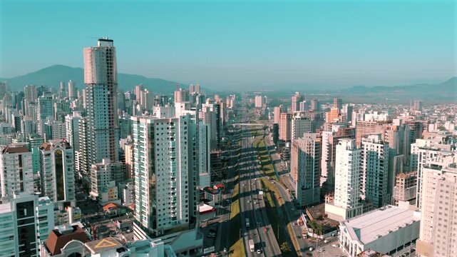 Avenida Nereu Ramos cutting through the skyline of Itapema, Brazil, with high-rise buildings, mountain backdrop, and clear skies representing Santa Catarina&rsquo;s urban coast, static drone shot