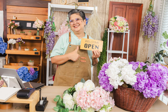 Asian senior woman, florist, small business owner proudly holds open sign with smile, ready to welcome customers to flower shop, concept of new beginning, retail startup, elderly with passion success