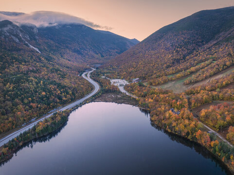 Aerial view of the winding road mirroring the serene lake's edge, nestled between mountains ablaze with autumn colors, Franconia Notch State Park, Vermont, United States.