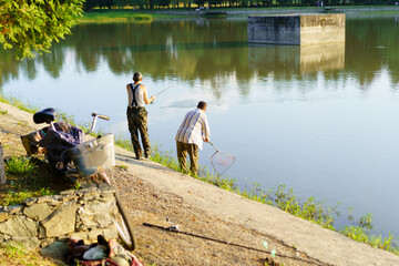 Two fishermen casting lines during sunset at a tranquil lake surrounded by greenery in a rural...