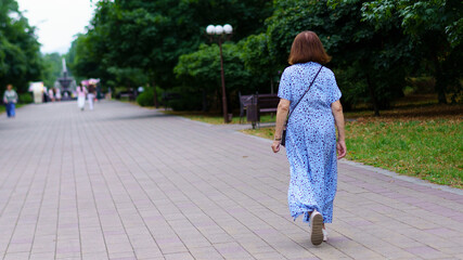 Woman in blue dress walking alone on a tranquil park path surrounded by greenery in warm afternoon...