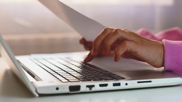 A close-up shot of hands typing on a white laptop keyboard while holding a document or paper, indicating a person working or studying.
