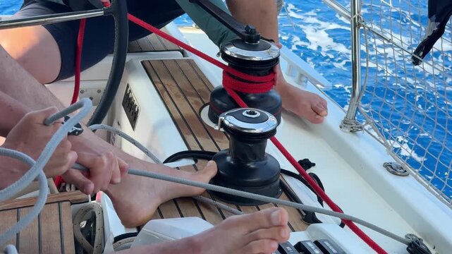 Close-up of hands tightening rope on winch during sail hoisting on sailing yacht. Teamwork and manual control in recreational boating activity.