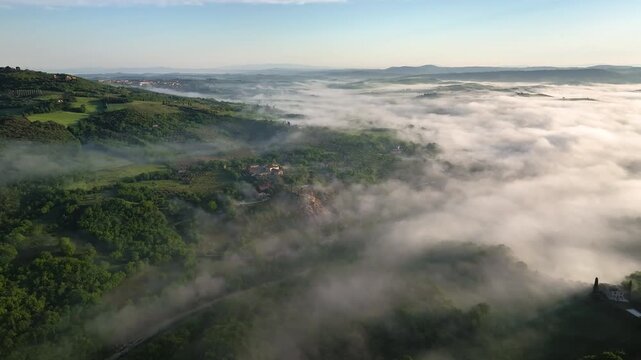 Aerial view of rolling green hills and the small town of Bagno Vignoni, partially obscured by a blanket of thick fog, creating a serene and ethereal landscape, Tuscany, Italy.