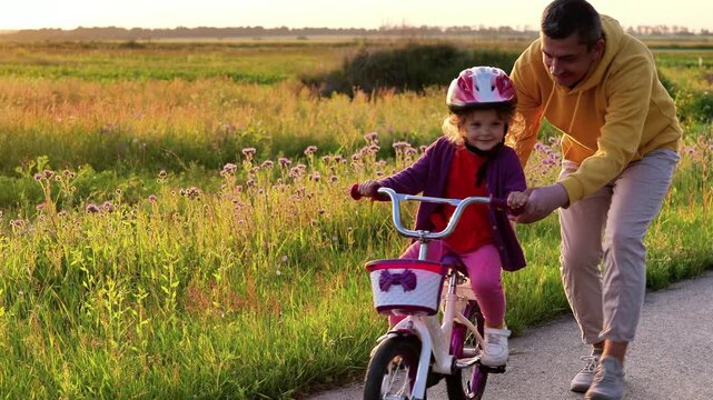 A father supports his young child who is wearing a red helmet and a purple cardigan while learning to ride a bicycle on a paved path next to a golden, grassy field at sunset.