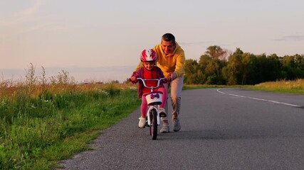 A close-up of a father helping his young child ride a bicycle on a paved road, with a field of green grass and trees in the background during golden hour. - Powered by Adobe