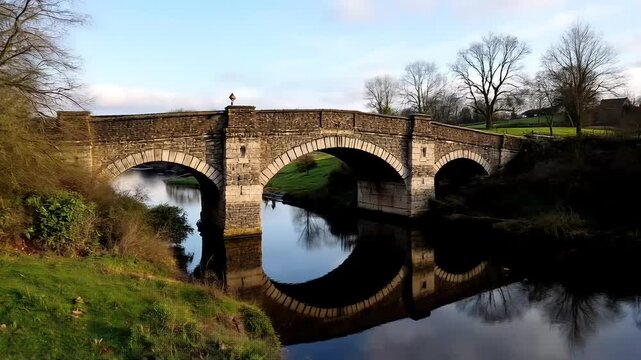 Ancient stone arch bridge over a European river, reflecting historic architecture and the surrounding natural landscape under an expansive sky High-quality cinematic natural 4k video