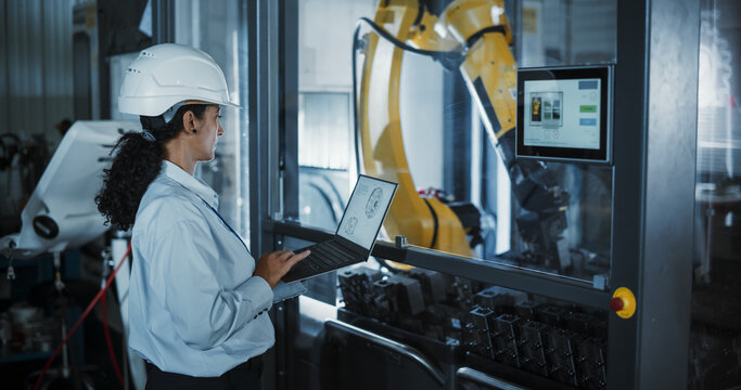 Young Hispanic Female Technician in a White Hard Hat Monitoring Manufacturing Process at an Advanced Metal Components Plant. Industrial Engineer Using Laptop Computer to Automate a Robotic Arm