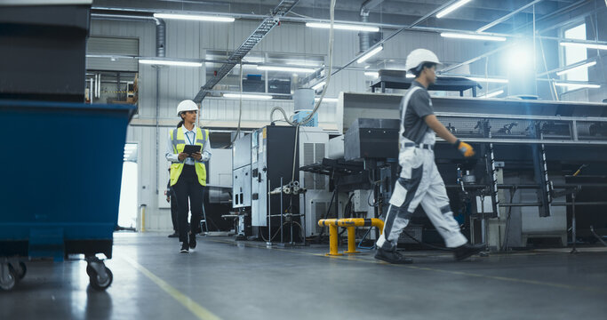 Hispanic Female Worker in a Reflective Vest Walks Through a Modern Factory, Inspecting Equipment and Recording Data on a Tablet Computer. Employees Working at the Facility with Advanced Machinery - Powered by Adobe