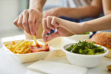 Unrecognizable couple eating together fast food, close-up cropped view, two hands dipping French fries into ketchup, burger, bowl of salad on table. Shared meals, food delivery, take-out food concept