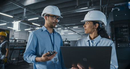 Two Multiethnic Employees Having a Conversation in an Industrial Factory Hall. Female Engineer Using a Laptop, Explaining the Scope of Work to a Young Technician. Workers Wearing Hard Hats for Safety