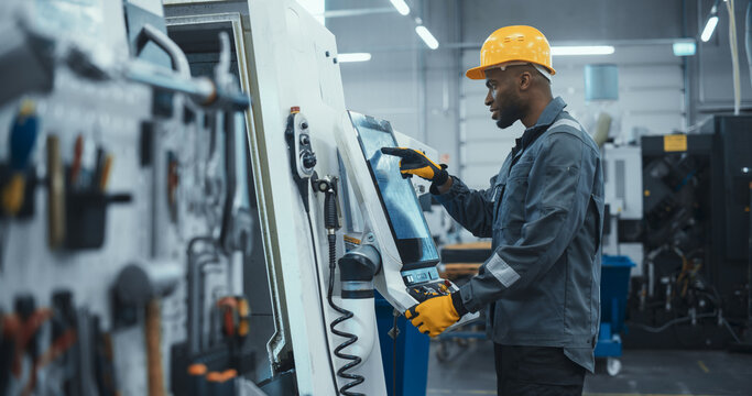 African Male Engineer in a Yellow Hard Hat Operating a CNC Milling Machine in a Modern Factory. Young Technician Inserting Parameters on a Touch Screen Display, Automating Production Line - Powered by Adobe