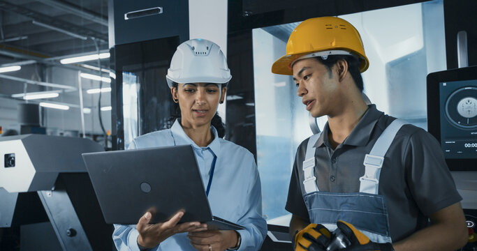 Two Male and a Female Factory Employees Standing in a High Tech Manufacturing Facility, Discussing Operations and Production Machinery. Diverse Employees Wear Safety Gear and Use a Laptop Computer
