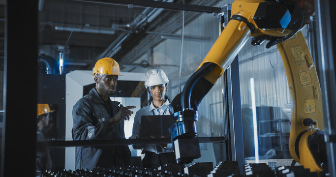 Industrial Manufacturing Facility: Young Black and Hispanic Engineers Controlling a Robotic Arm with a Tablet Computer App. Operators Downloading New Production Instructions for the Machine