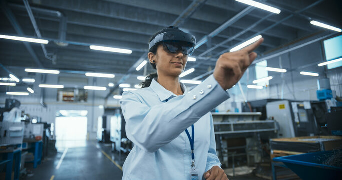 Multiethnic Female Engineer in a Mixed Reality Headset Standing in a High Tech Manufacturing Facility, Using Modern Technology to Operate Precision Engineering Tools at a Factory