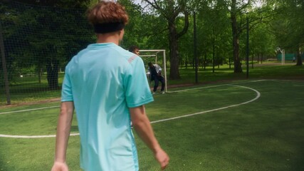 Slowmo of teen soccer player practicing kicking ball toward goalpost while coach standing in goal and two teammates nearby during outdoor training on sunny field
