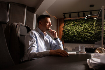 Focused man in a light shirt holds smartphone while working at a stylish evening office. Modern...