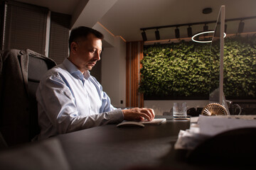 Focused man in a light shirt holds smartphone while working at a stylish evening office. Modern...