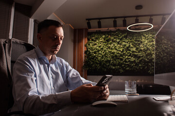 Businessman in a light shirt types on keyboard and phone in a dark elegant office. Evening light,...