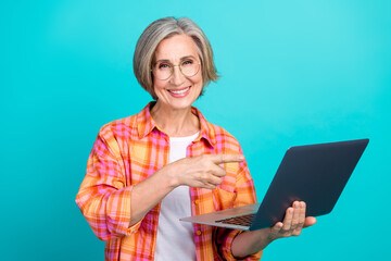 Smiling elderly woman holding a laptop pointing at the screen against a vibrant turquoise...