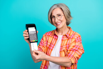 Smiling elderly woman in casual checkered shirt presenting a credit card and payment terminal...