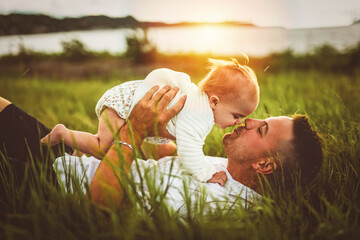 father and baby child girl at the sunset lay on the grass on the waterscape view from back